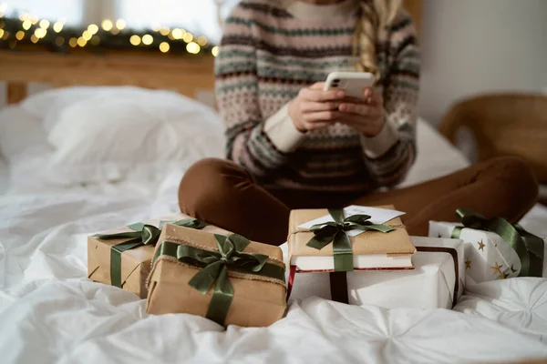 无法辨认的高加索女人坐在圣诞礼物中 一边躺在床上一边用电话 — 图库照片