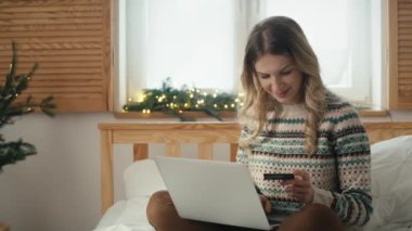 Neşeli beyaz kadın yatakta oturuyor ve Noel alışverişi yapıyor dizüstü bilgisayar ve kredi kartıyla. 8K 'da kırmızı helyum kamerayla çekildi..      