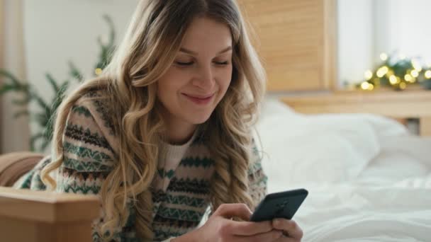 クリスマスの時間にベッドの前に横になって電話を使っている白人女性 8Kでレッドヘリウムカメラで撮影 — ストック動画