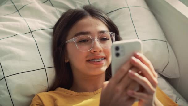 彼女の背中と笑顔にベッドの上に横たわっている間 白人の十代の少女は電話をブラウジング 8Kでレッドヘリウムカメラで撮影 — ストック動画
