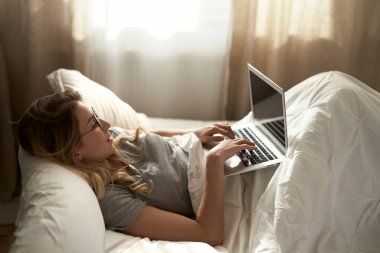 Kafkasyalı kadın dizüstü bilgisayar kullanıyor ve sabahları yatakta yatıyor.