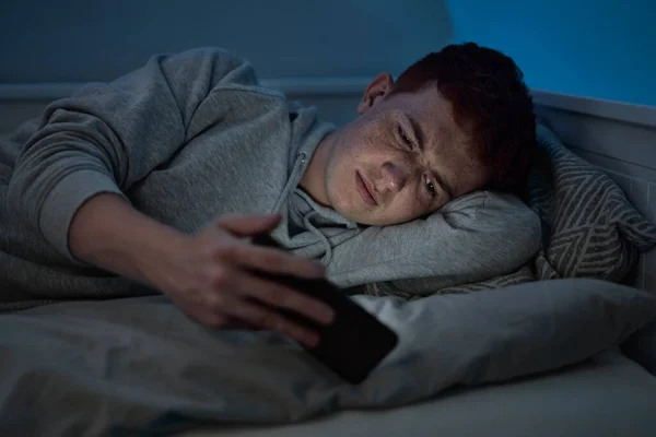 夜にベッドの上に横たわっている間白人の十代の男の子のブラウジング電話 — ストック写真
