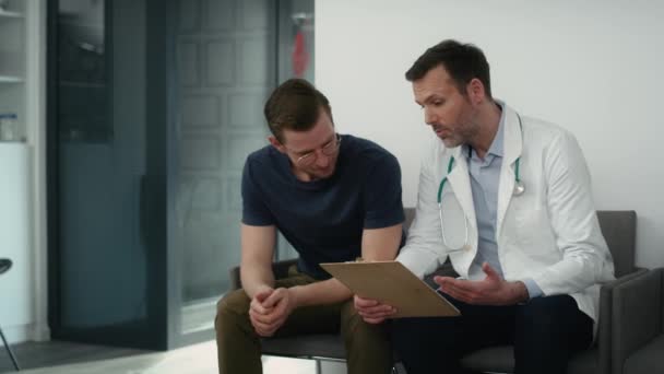 白人男性医師と患者は保健所の廊下で文書を議論座っている 8Kでレッドヘリウムカメラで撮影 — ストック動画