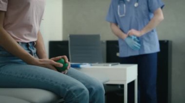 Doktor arka plandaki tıbbi muayeneye hazırlanırken beyaz bir kadın tıp yatağında oturmuş stres topu sıkıyor. 8K 'da kırmızı helyum kamerayla çekildi.. 