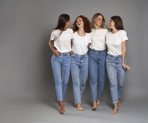 四个年轻的高加索女人穿着蓝色牛仔裤和灰色背景的白衬衫走路 — 图库照片