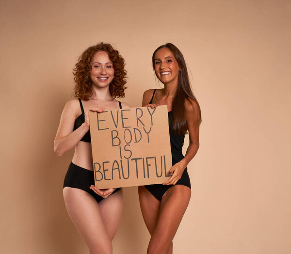 Two caucasian women in underwear standing on nude background in studio and holding banner