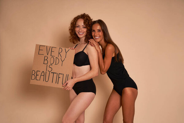 Two caucasian women in underwear standing on nude background in studio and holding banner 