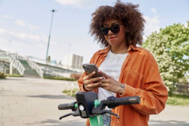 Genç siyahi kadın elektrikli scooter kullanmak için cep telefonu kullanıyor.