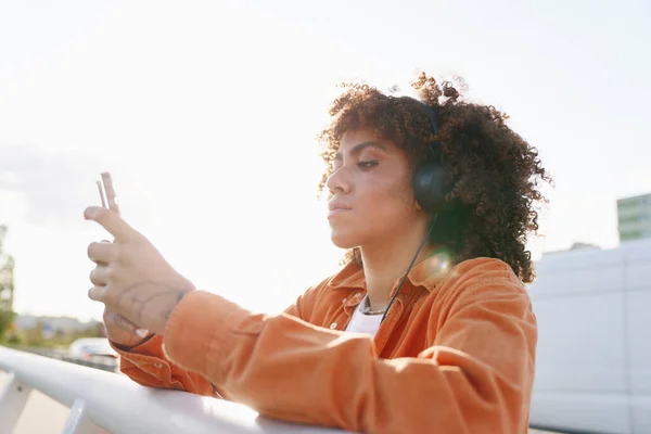 橋の上に立っている間にヘッドフォンや携帯電話を身に着けている黒人女性 — ストック写真