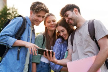 Üniversite öğrencileri dışarıda duruyor ve cep telefonlarına bakıyorlar 