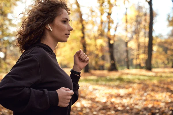 秋天穿着耳机在公园里慢跑的高加索女人的中间镜头 — 图库照片