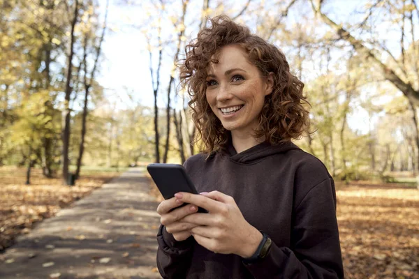 白种人女人一边浏览手机一边望着秋天的公园 — 图库照片