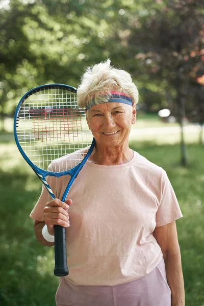 Portrait of senior woman standing in the park and holding tennis racket