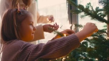 Neşeli anne ve kız Noel ağacını ışıklarla süslüyorlar. 8K 'da kırmızı helyum kamerayla çekildi..   