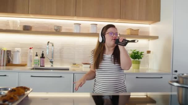 エネルギッシュダウン症候群の女性は キッチンでヘッドフォンとダンスを身に着けています 8KでRedヘリウムカメラで撮影する — ストック動画