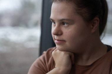 Düşünceli Down sendromlu kadın pencerenin yanında oturuyor ve başka tarafa bakıyor.