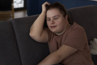 Düşünceli Down sendromlu kadın oturma odasında oturuyor ve başka tarafa bakıyor.