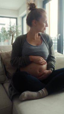Kafkasyalı hamile bir kadının kanepeye oturup karnına dokunduğu dikey video.