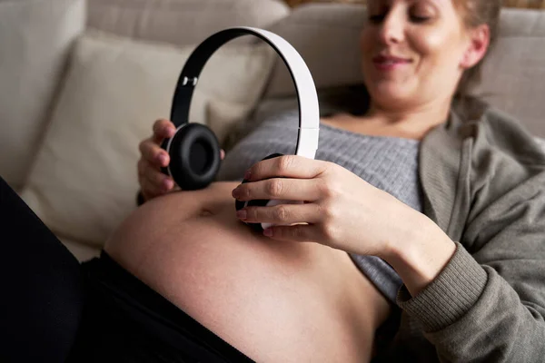 Biała Kobieta Ciąży Leżąca Kanapie Zakładająca Słuchawki Brzuch — Zdjęcie stockowe