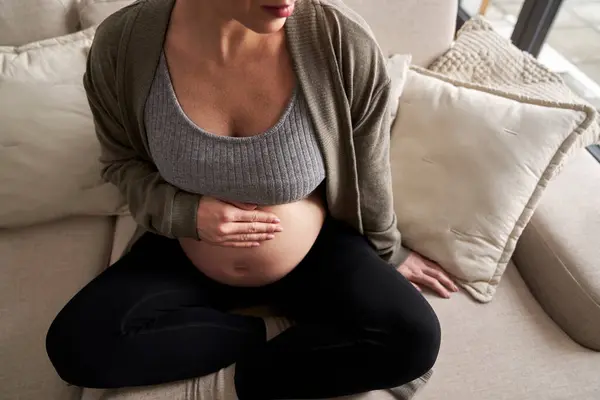 无法辨认的高加索孕妇坐在沙发上摸着她的腹部 — 图库照片