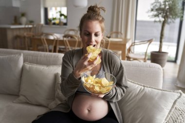 Hamile kadın koltukta otururken cips yiyor.