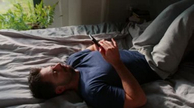 Sıkılmış adam yatağa uzanıp cep telefonuna bakıyor.