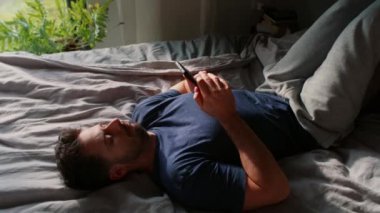Sıkılmış adam yatağa uzanıp cep telefonuna bakıyor.