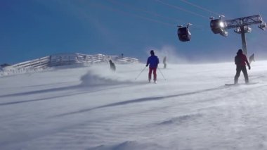 Slovakya. Kayak merkezindeki güneşli kış günü Jasna ve yamaçtaki kar fırtınası. Kayakçılar kayak asansörünün arka planına inerler. Yavaş çekim