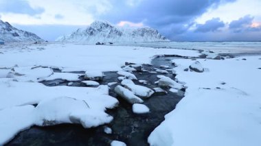 Kış Norveç. Lofoten Adaları. Dere, dalgaların ve karla kaplı dağların arka planına karşı bir kış plajında okyanusa akar.