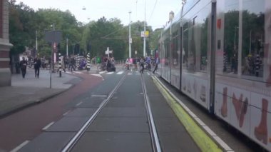 Hollanda, Amsterdam - 27 Eylül 2021: Gündüz. Şehir merkezindeki yeşil bir parka giden bir tramvaydan yaya geçidinde yaya ve bisikletçiler görülüyor. Yavaş çekim