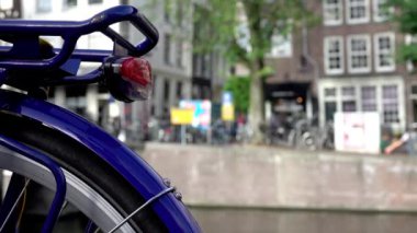 Hollanda 'da. Amsterdam Kanalı 'nın setinde yaz sabahı. Bisikletin arka tekerine yakın çekim. Arka planda bisikletlerin, arabaların ve insanların odaksız hareketi