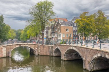 Hollanda, Amsterdam - 26 Eylül 2021: Bulutlu bir yaz günü. Amsterdam kanallarındaki eski taş köprüler. Toprak setin üzerinde dans eden tipik Hollandalı evleri. Mobiletli gençler.