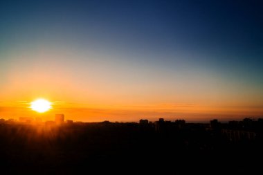 Şehir üzerinde açık bir yaz gökyüzünde şafak. Yükselen güneş ufuktaki ışık bulutlarını delip geçer. Güneş ışınlarının sisi binaların siluetlerine karşı