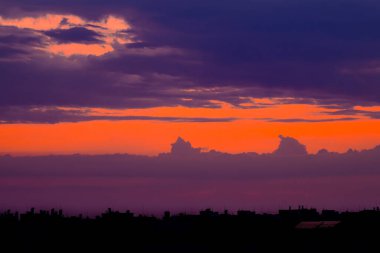 Şehrin çatılarında bulutlu gökyüzü. Güneş, binaların siluetlerinin arkasında kayboldu. Gökyüzünü kırmızı ve turuncu tonlarda boyuyor.