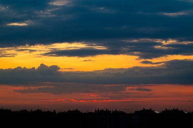 Şehrin çatılarının üstündeki bulutlu gökyüzü. Güneş, binaların siluetlerinin arkasına saklanmıştı. Gökyüzünü güzel renklerle boyamıştı.