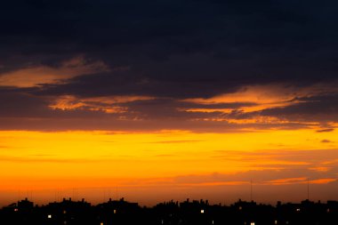 Şehrin çatılarında bulutlu bir gökyüzü. Güneş, binaların siluetlerinin arkasına zar zor gizlenmiş, gökyüzünü güzelce resmediyor. Evlerin pencerelerinde ışıklar yanıyor.