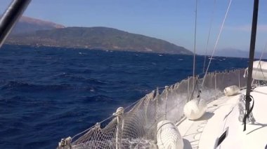 Güneşli ve rüzgarlı bir gün. Beyaz yelkenli yat kıyıya yakın deniz dalgaları üzerinde yelken açar ve bir su püskürtme bulutu yaratır. Yavaş çekim