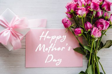 Happy mother's day text sign. Postcard with bouquet of small pink roses, gift box on white rustic wooden background. Greeting card with flowers concept. Holiday greeting card! Top view, flat lay.