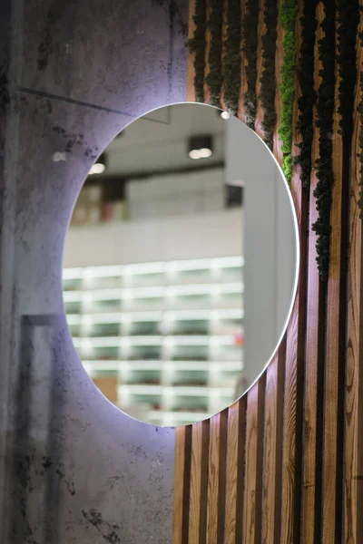 A large round mirror with a reflection of the room. A mirror hangs on a concrete wall and a wooden background with green moss, and greenery in the light living room. Modern interior details. Closeup.