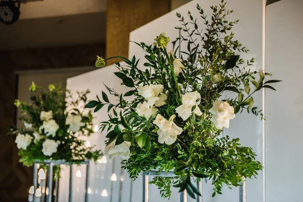 Decor of flowers and greenery for a wedding ceremony in a restaurant. The metal arch is decorated white and green flowers and garlands with light bulbs. Details interior closeup.