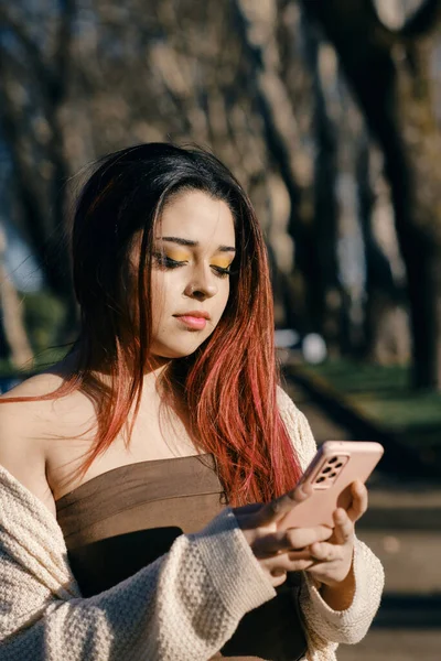 Red-haired young Latina woman chats with her cell phone in outdoor park