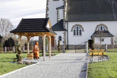 A joyful figure of Pope John Paul II made of wood by a folk artist. The Pope is sitting on a bench under a wooden roof. clipart