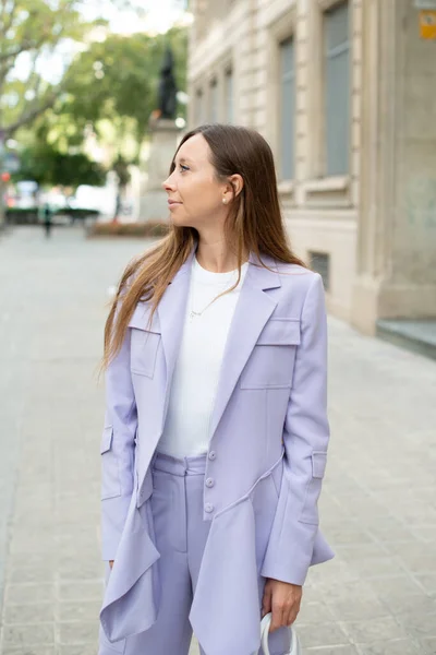 Street style portrait of a beautiful woman walking in the city center. Stylish happy young woman standing in the street in trendy lilac outfit. Spring season. Selective focus photo