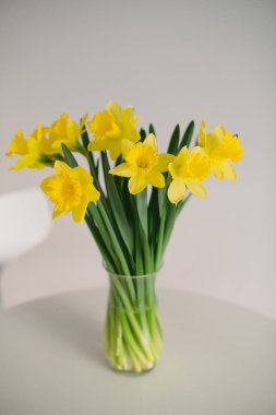 A bouquet of daffodils in a glass vase on a white table against a white wall, front view, copy space. Decorating home with spring flowers. Selective focus photo.