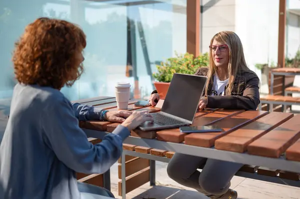 İki iş kadını açık bir kafede dizüstü bilgisayarları ve tabletleri üzerinde çalışıyor, kahve molası veriyor ve iş konularını tartışıyorlar.