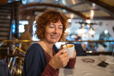 Büyüleyici bir restoranda rahatlatıcı bir anda cappuccinonun tadını çıkaran gülümseyen olgun bir kadın.