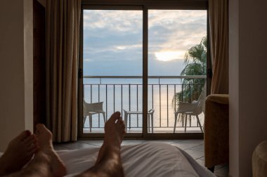 Çift bacakları yatakta dinleniyor, otel odasının balkonundan okyanusun üzerinde güzel bir güneşin doğuşunun tadını çıkarıyor.