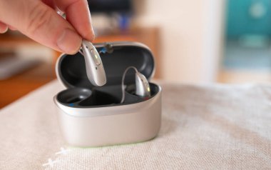 Fingers holding a hearing aid with its charging and storage case on a table, highlighting accessibility and convenience for hearing impaired individuals clipart