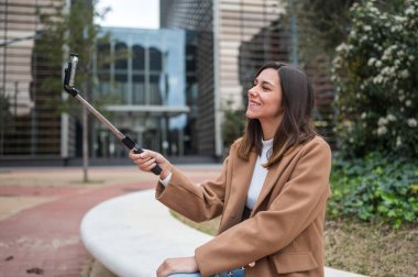 Gülümseyen iş kadını, şehir parkında uzatma çubuğu kullanarak selfie çekiyor.