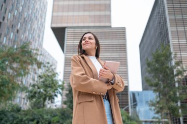 İş kadını elinde tabletle modern bir şehrin gökdelenlerine bakıyor ve başarılı kariyerinin tadını çıkarıyor.